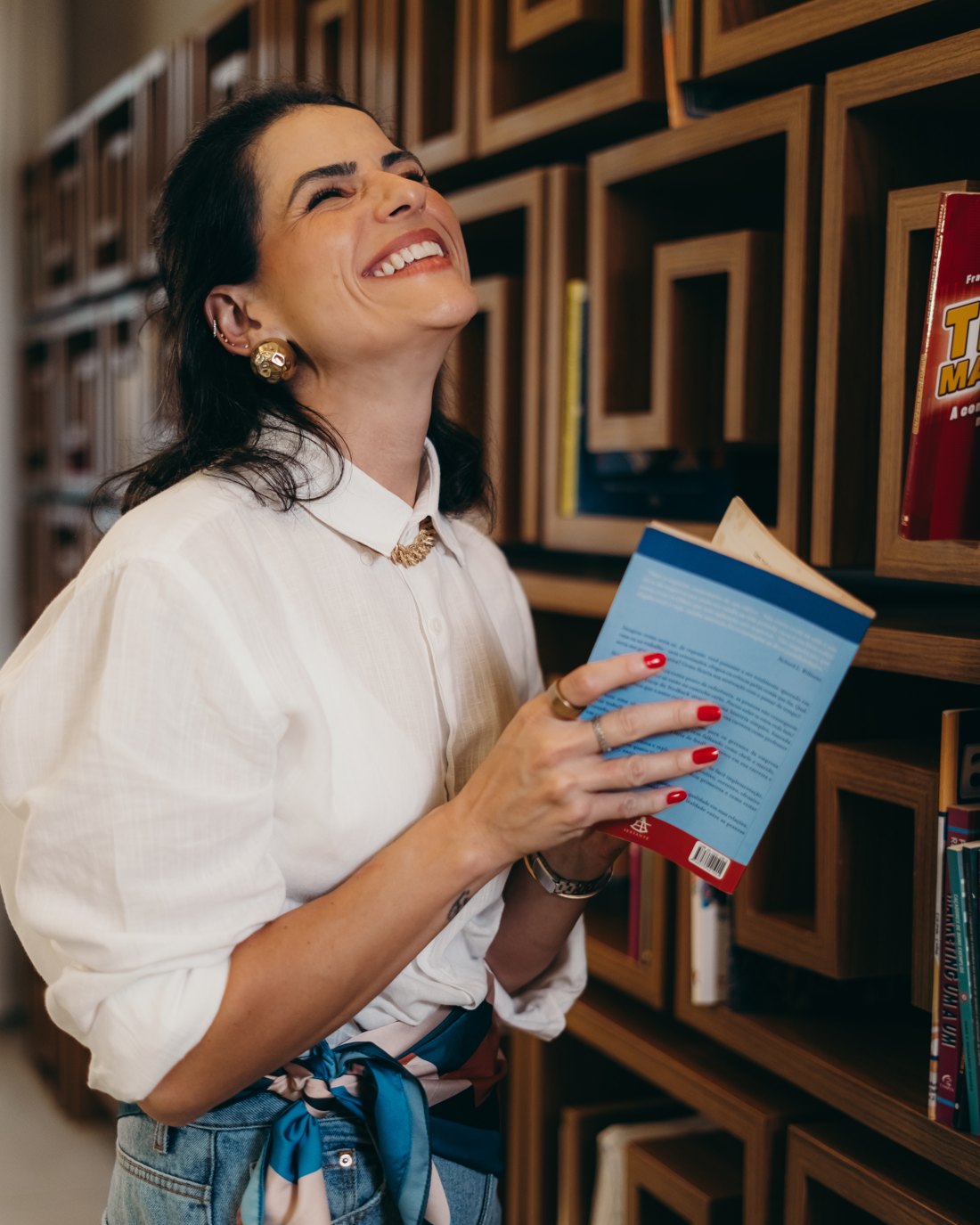 Mariana Leite, sorrindo, segurando um livro em frente a uma estante.